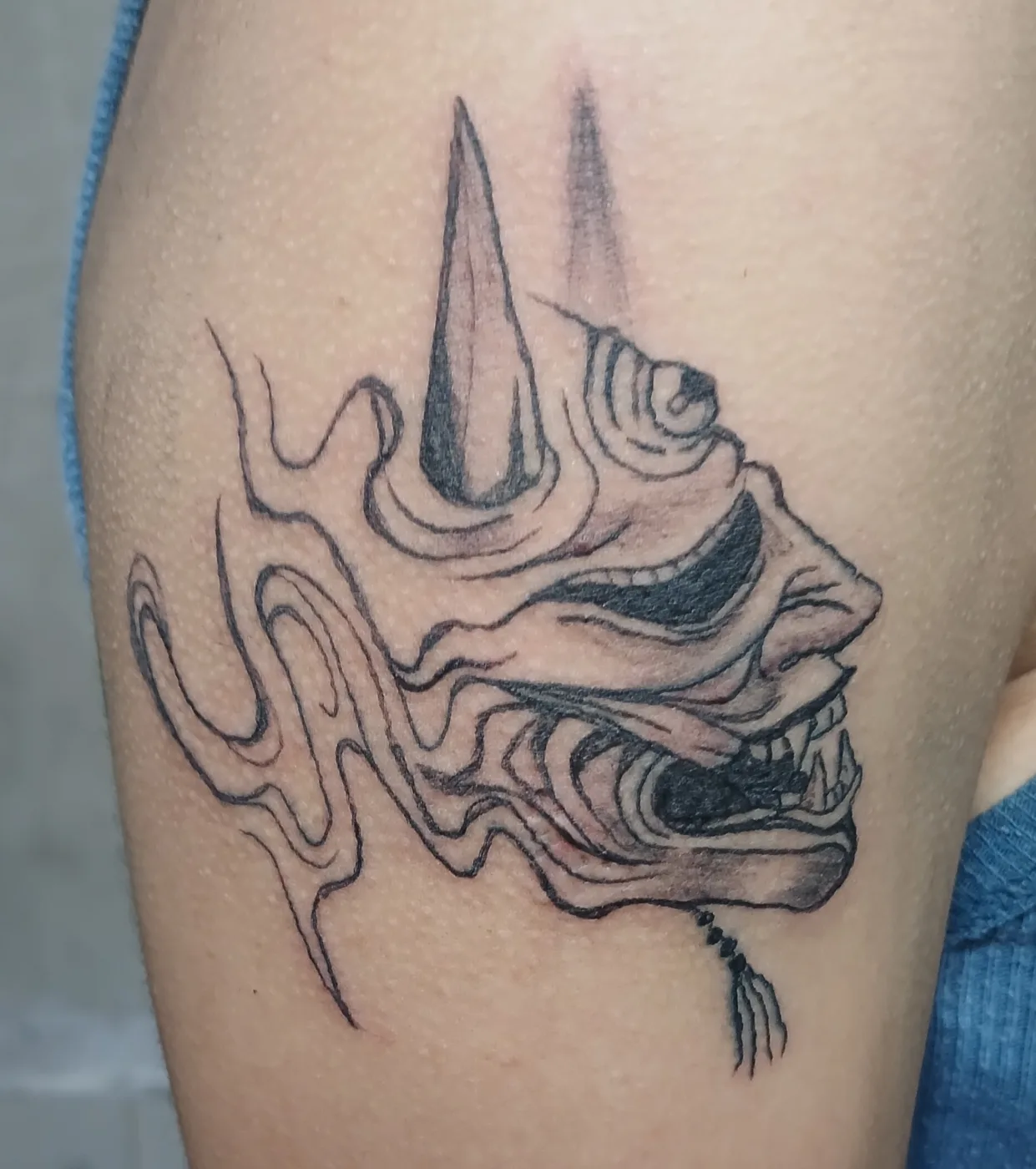 Máscara oni entre humo, tatuada sobre el brazo con línea limpia.