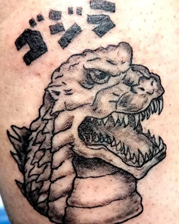 Cabeza de monstruo tipo kaiju con letras japonesas, tatuada en piel con línea y sombreado.