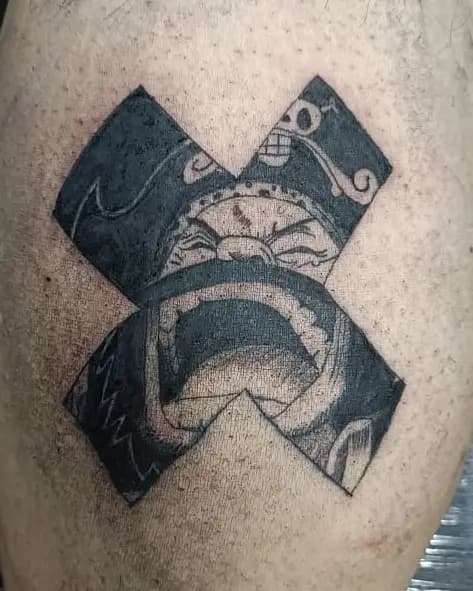 Una X en negro sólido con un rostro ornamentado en su interior, tatuada en piel.