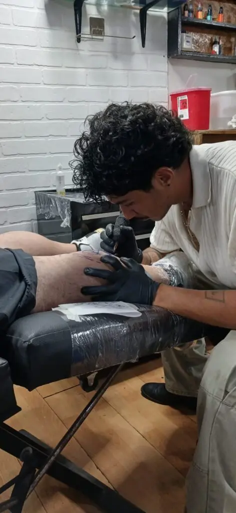 Alumno tatuando la pierna de una persona en el estudio, con guantes negros y la camilla cubierta con plástico protector.